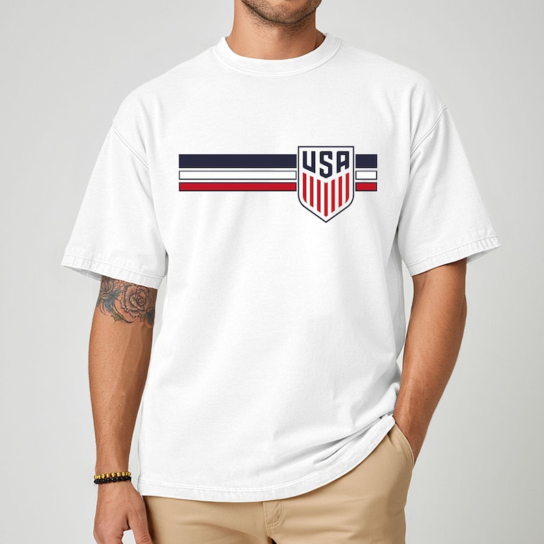 Puede incluir: Una camiseta blanca de manga corta con un dise&ntilde;o gr&aacute;fico horizontal en el pecho. El dise&ntilde;o consta de tres franjas paralelas en azul marino, blanco y rojo, cruzadas por un emblema en forma de escudo que contiene el texto USA en color azul.