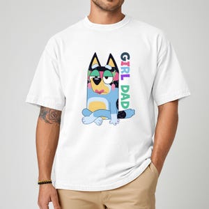 May include: White t-shirt featuring a cartoon illustration of a blue dog with floral accents and the text "GIRL DAD" in colorful letters. The dog is sitting with its legs crossed. The shirt has short sleeves.