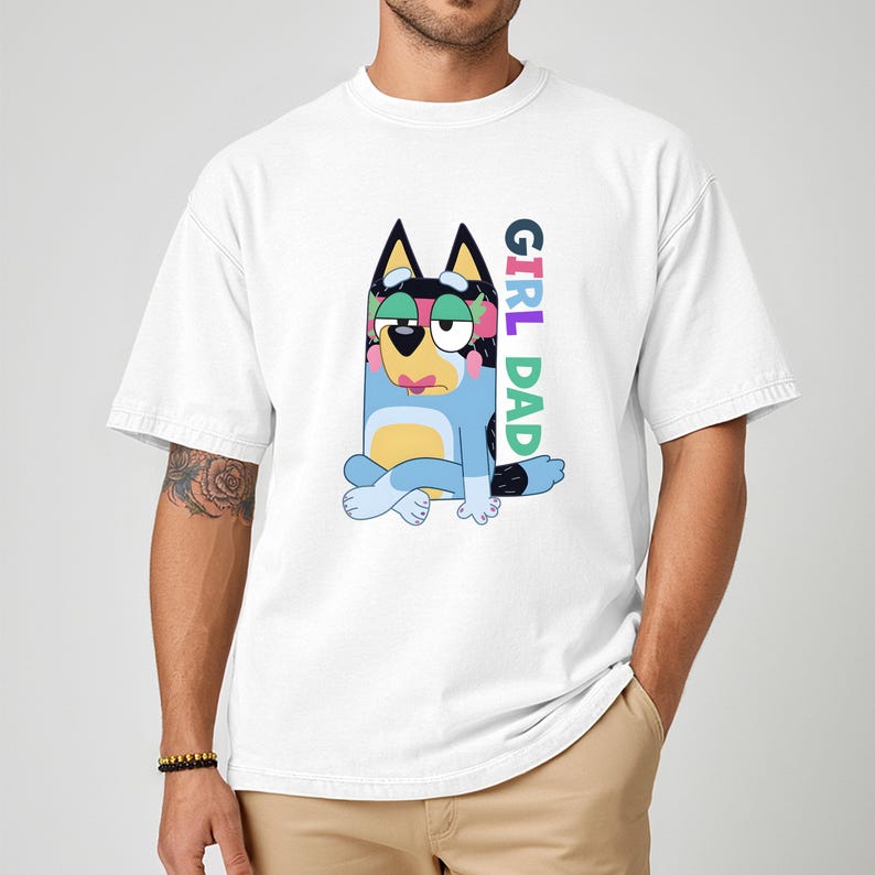 May include: White t-shirt featuring a cartoon illustration of a blue dog with floral accents and the text "GIRL DAD" in colorful letters. The dog is sitting with its legs crossed. The shirt has short sleeves.