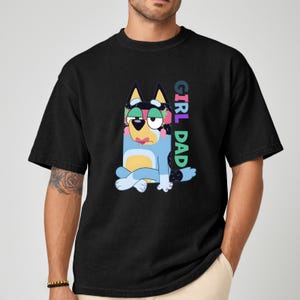 May include: Black t-shirt featuring a cartoon dog character in blue, yellow, and black with the text "GIRL DAD" in colorful letters. The dog is sitting with its legs crossed. The shirt has short sleeves and a crew neck.
