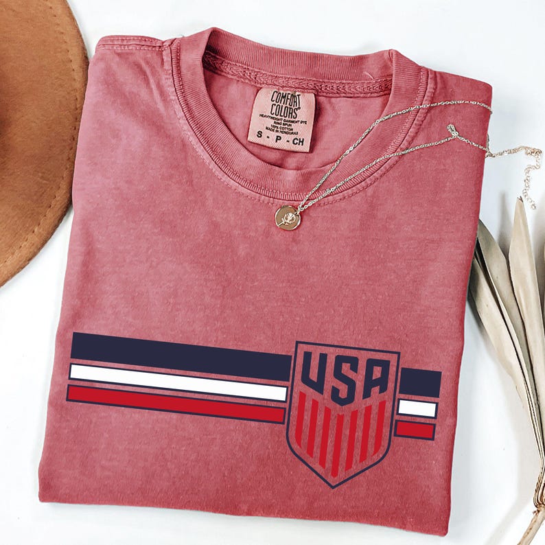 Puede incluir: Una camiseta de cuello redondo en color rosa empolvado con un dise&ntilde;o gr&aacute;fico de tres franjas horizontales en azul marino, blanco y rojo. A la derecha, un emblema en forma de escudo contiene el texto USA sobre franjas verticales rojas.