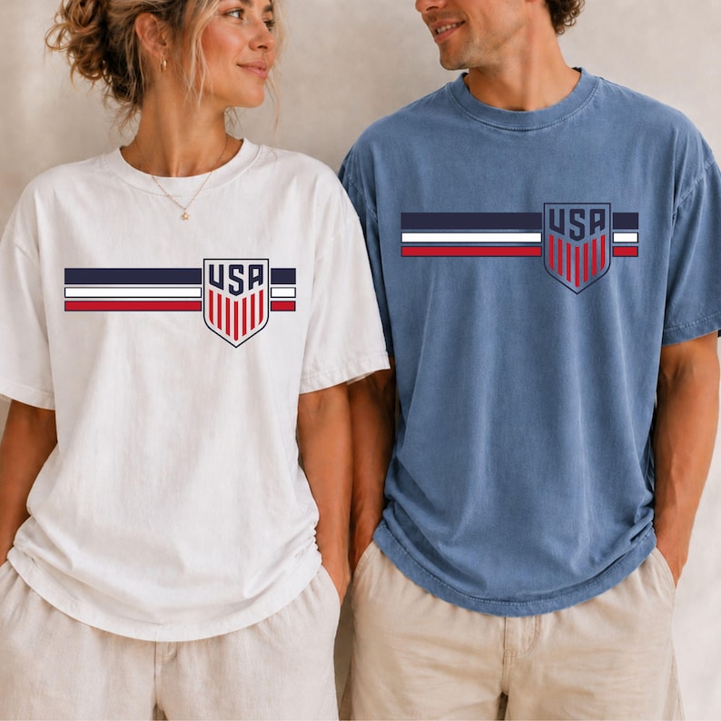 Puede incluir: Una camiseta blanca y una camiseta azul, cada una con un dise&ntilde;o de rayas horizontales en azul marino, blanco y rojo en el pecho. Un emblema en forma de escudo est&aacute; centrado sobre las rayas, conteniendo el texto USA sobre rayas verticales.