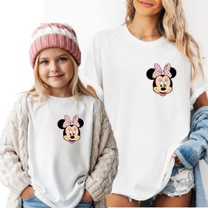 Op de afbeelding: Witte T-shirts met een Minnie Mouse-afbeelding met een roze stippenstrik. E&eacute;n shirt wordt gedragen door een kind met een roze en witte gestreepte muts en een cr&egrave;mekleurig vest. Het andere shirt wordt door een volwassene gedragen.