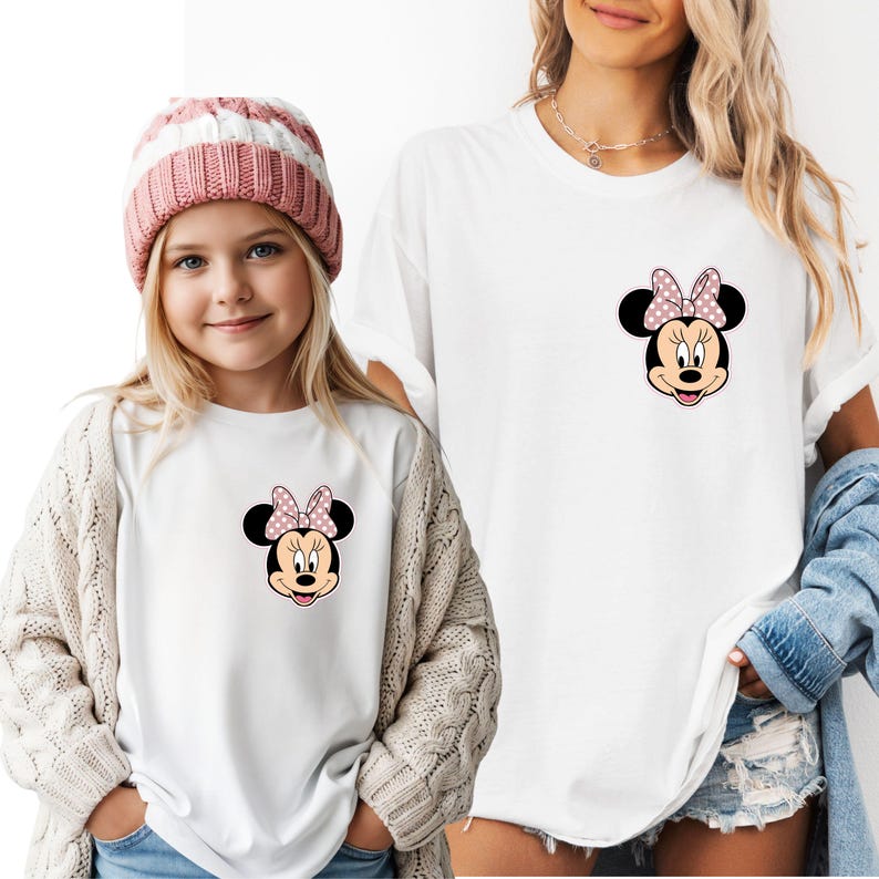 Op de afbeelding: Witte T-shirts met een Minnie Mouse-afbeelding met een roze stippenstrik. E&eacute;n shirt wordt gedragen door een kind met een roze en witte gestreepte muts en een cr&egrave;mekleurig vest. Het andere shirt wordt door een volwassene gedragen.