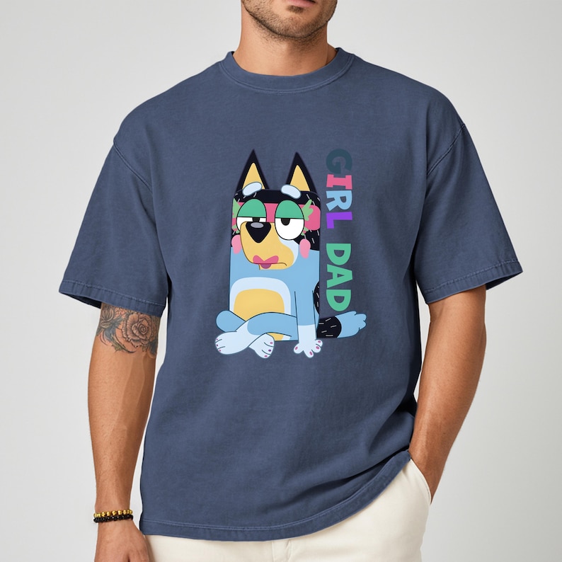 May include: A navy blue t-shirt featuring a cartoon dog character with the text "GIRL DAD" in vertical, multicolored letters. The dog is blue, yellow, and black, with a sad expression. The shirt has short sleeves and a relaxed fit.