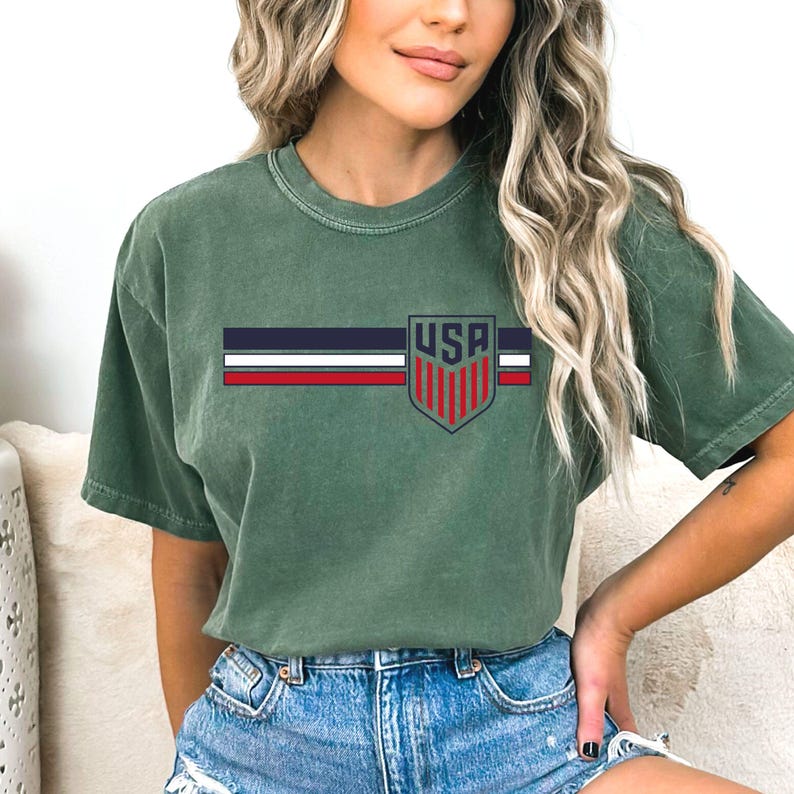 Puede incluir: Una camiseta de manga corta color verde salvia con un dise&ntilde;o gr&aacute;fico horizontal. El dise&ntilde;o consta de tres franjas paralelas en azul marino, blanco y rojo, cruzadas por un emblema en forma de escudo con el texto USA sobre rayas rojas verticales.