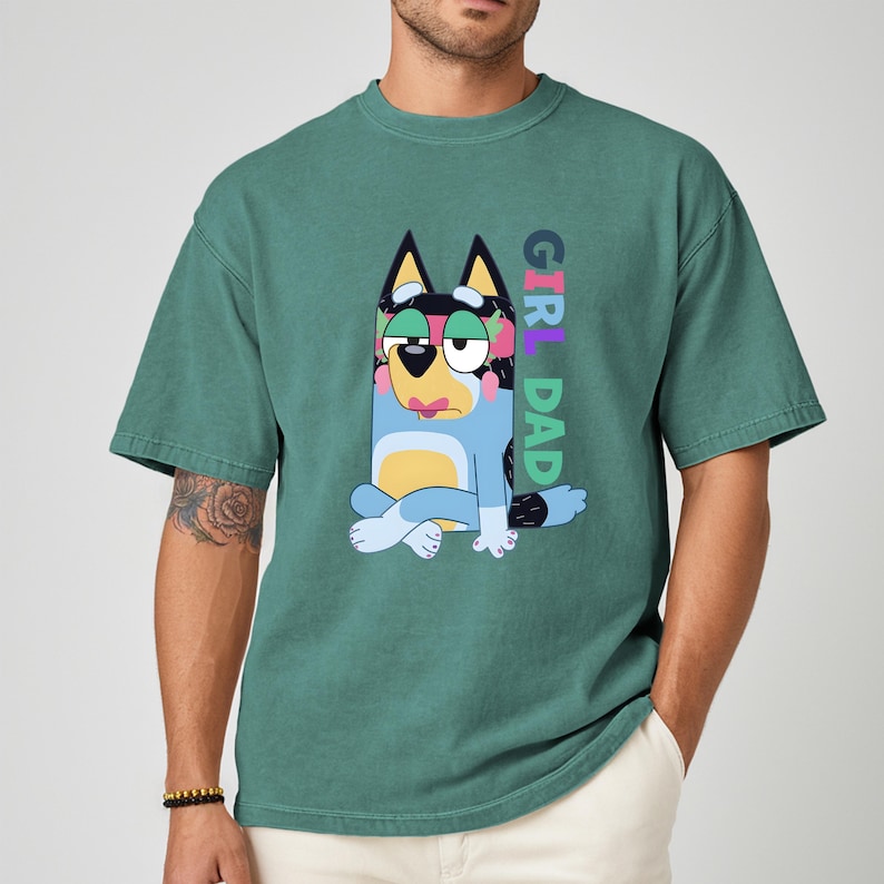 May include: Teal green t-shirt featuring a cartoon dog character with the text "GIRL DAD" in colorful letters. The dog is blue with black ears and a pink tongue. The shirt has a relaxed fit.