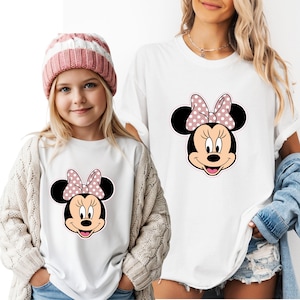May include: White t-shirts with a Minnie Mouse design. The design shows a black head, ears, and a pink bow with white polka dots. One shirt is worn by a child wearing a pink and white striped hat and a cream cardigan.