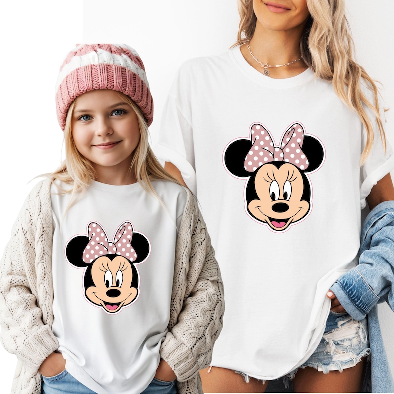 May include: White t-shirts with a Minnie Mouse design. The design shows a black head, ears, and a pink bow with white polka dots. One shirt is worn by a child wearing a pink and white striped hat and a cream cardigan.
