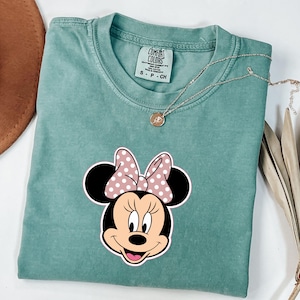 May include: A seafoam green t-shirt with a Minnie Mouse graphic, featuring a pink polka dot bow. The shirt has a Comfort Colors tag and a silver necklace with a pendant. The shirt is folded on a white surface.