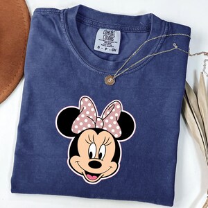 May include: A navy blue Comfort Colors t-shirt with a Minnie Mouse graphic. The graphic shows Minnie's face with a pink polka dot bow. A gold necklace with a pendant rests on the shirt. The shirt is folded neatly.