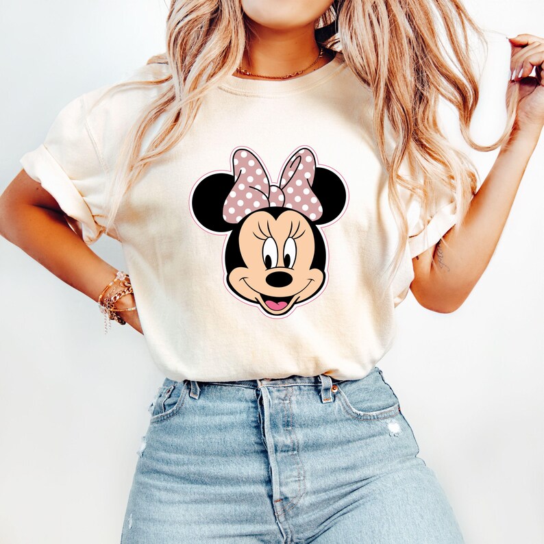 May include: A cream-coloured t-shirt with a Minnie Mouse graphic. The graphic depicts Minnie's face with black ears, a pink bow with white polka dots, and a pink outline. The shirt is worn with blue jeans.