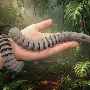 Puede incluir: Un juguete de serpiente articulada gris con un diseño segmentado detallado. El juguete se sostiene en una mano sobre un exuberante fondo de selva verde. La cabeza de la serpiente presenta detalles realistas de los ojos.