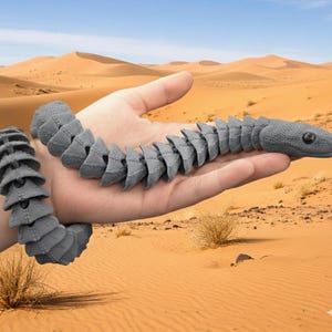 Puede incluir: Un juguete de serpiente articulada gris, sostenido en una mano, con un cuerpo segmentado y una cabeza detallada. El juguete está hecho de un material texturizado. El fondo presenta un paisaje desértico con dunas de arena.