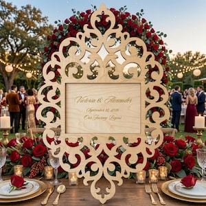 Puede incluir: Un letrero de boda de madera con un diseño calado ornamental. El letrero presenta los nombres "Victoria & Alexander" y la fecha "12 de septiembre de 2026". Rosas rojas y velas decoran la mesa. Un elemento decorativo para una boda.