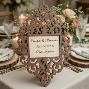 May include: A wooden wedding table sign with an ornate, openwork frame. The sign displays the names "Charlotte & Maximilian", the date "June 14, 2026", and the phrase "Better Together". The sign is set on a table with floral arrangements.