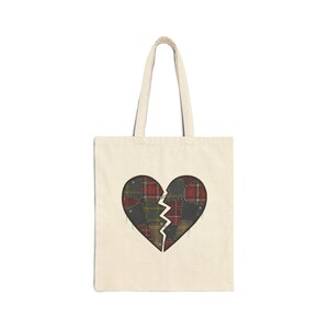 May include: Cream-coloured tote bag with a broken heart design. The heart is made of patchwork plaid fabric in red, green, and black. The bag has a simple design with long handles, ideal for carrying items.