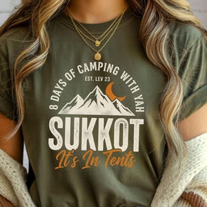 Może przedstawiać: Zielona koszulka z nadrukiem. Projekt zawiera tekst "8 Days of Camping with Yah" i "Sukkot It's In Tents". Grafika przedstawia również góry i półksiężyc.