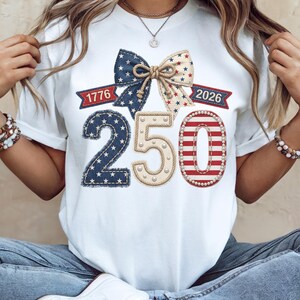 Puede incluir: Una camiseta blanca con un gráfico de un lazo grande con estrellas y rayas, los números 250 y los años 1776 y 2026. El diseño incorpora texturas de mezclilla, detalles de cuerda y adornos tipo perla en colores rojo, blanco y azul.