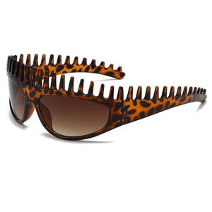 Puede incluir: Gafas de sol con un patrón de estampado de leopardo marrón y negro en la montura. La montura presenta un diseño distintivo con un borde superior dentado en forma de peine. Las lentes están tintadas en un tono marrón degradado más oscuro.