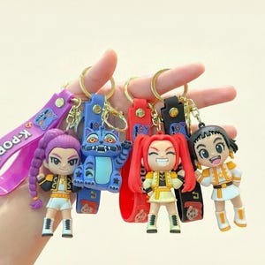 Może przedstawiać: Kolekcja breloków w stylu K-Pop z postaciami z kreskówek i niebieskim kotem. Breloki mają złote kółka i kolorowe paski z napisem "K-POP". Postacie noszą stroje w kolorze żółtym, białym i czarnym.