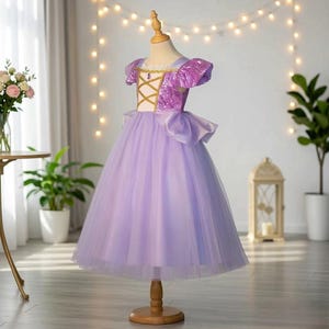 Op de afbeelding: Een lavendelkleurige prinsessenjurk op een mannequin. De lijfje heeft een gouden kruisontwerp en paillettenmouwen. De rok is van gelaagde tule en een grote strik accentueert de taille. De jurk is ontworpen voor een kind.