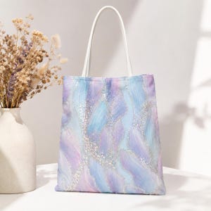 May include: A tote bag with a white handle and a colorful design. The bag features a pattern of flowing blue, purple, and white brushstrokes with sparkling accents. The bag is made of a soft material and is suitable for carrying various items.