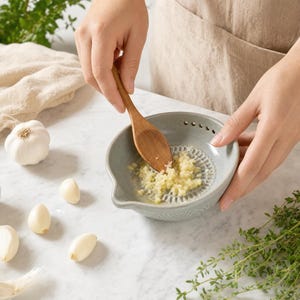 Puede incluir: Un cuenco rallador de ajo de cerámica gris claro con un centro texturizado y un pequeño pico vertedor, que contiene ajo picado. Se utiliza una pequeña cuchara de madera para raspar el ajo, mientras hay dientes de ajo y hierbas cerca.