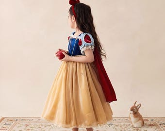 May include: A dress featuring a blue velvet bodice, puffed sleeves with red accents, and a long, layered gold tulle skirt. The outfit includes a red velvet headband with a bow and a long red cape attached to the back, while holding a red apple.