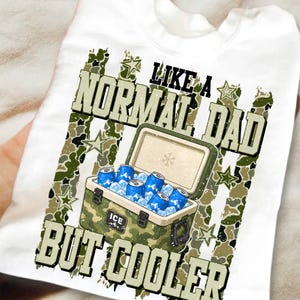 May include: White t-shirt with a graphic design featuring a camouflage cooler filled with blue cans and ice. The text reads "LIKE A NORMAL DAD BUT COOLER" in a bold, green font. The design includes star accents.