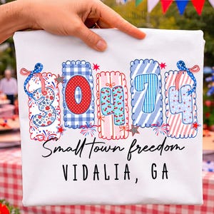May include: A white t-shirt featuring a graphic with the number 30474 in colorful, patterned, scalloped-edge blocks. Below the numbers, the text reads Small Town freedom and VIDALIA, GA in black script and sans-serif fonts respectively.
