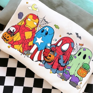 May include: A white sweatshirt featuring a graphic print of four characters dressed as ghosts for Halloween. The characters resemble Iron Man, Captain America, Spider-Man, and a green ghost, each carrying a jack-o'-lantern candy bucket.