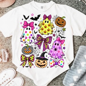 May include: A white t-shirt featuring a colorful Halloween-themed graphic print. The design includes three ghosts, three pumpkins, several patterned bows, spiderwebs, bats, and stars in shades of pink, yellow, orange, and black.