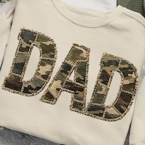 May include: Cream-colored sweatshirt with the word "DAD" in large, camouflage-patterned letters. The letters are outlined with a gold-colored stitching, creating a textured and eye-catching design. The sweatshirt has a classic crew neck.