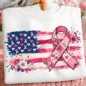 May include: A white sweatshirt featuring a graphic of a distressed American flag in shades of pink and white. The flag includes pink hearts and ribbons in the blue field, with a large pink floral-patterned awareness ribbon layered over the stripes.