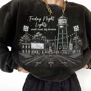 Op de afbeelding: Een zwart sweatshirt met ronde hals en een witte grafische illustratie van een voetbalveld, een watertoren met het opschrift Petersburg en stadionverlichting. De tekst luidt Friday Night Lights en small town. big dreams.