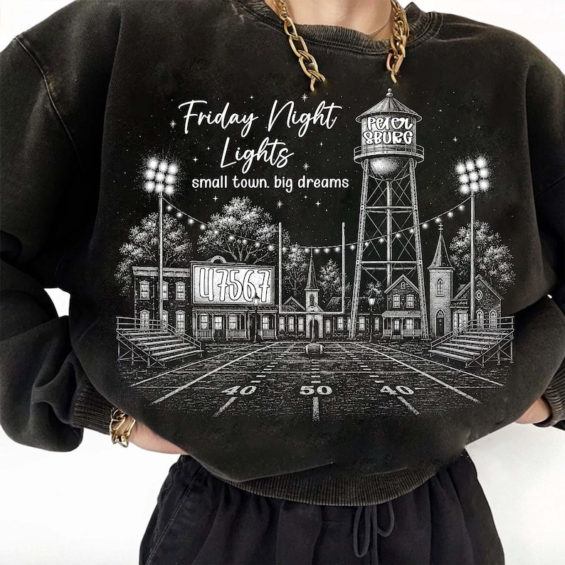 Op de afbeelding: Een zwart sweatshirt met ronde hals en een witte grafische illustratie van een voetbalveld, een watertoren met het opschrift Petersburg en stadionverlichting. De tekst luidt Friday Night Lights en small town. big dreams.