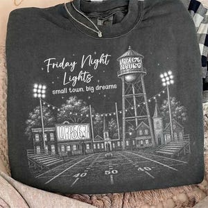 Op de afbeelding: Een antracietkleurige sweater met ronde hals en een witte grafische print van een voetbalveld, een watertoren met het opschrift Petersburg en stadionverlichting. De tekst luidt Friday Night Lights en small town. big dreams.