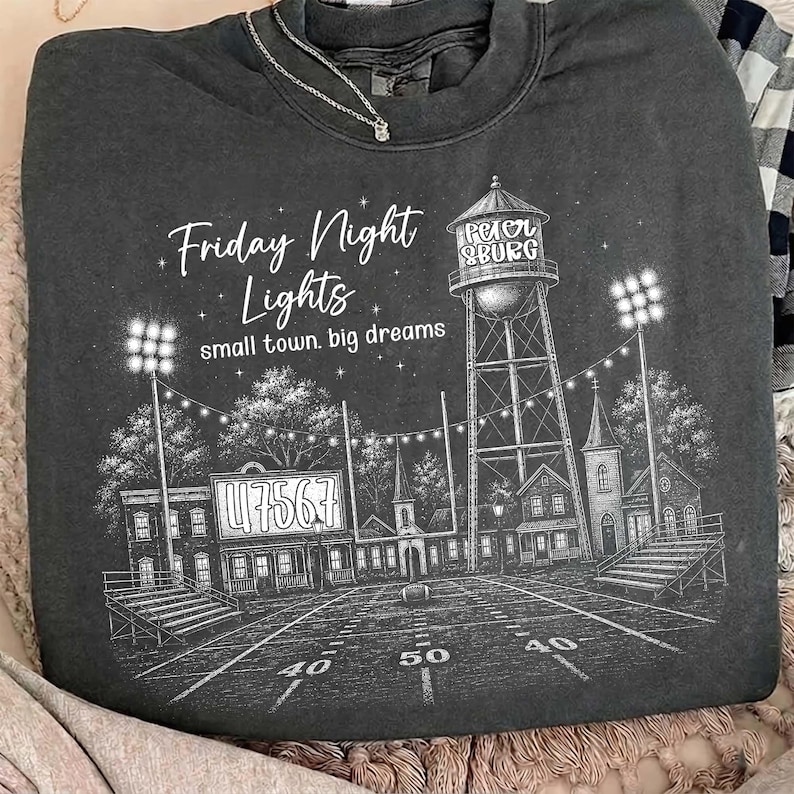 Op de afbeelding: Een antracietkleurige sweater met ronde hals en een witte grafische print van een voetbalveld, een watertoren met het opschrift Petersburg en stadionverlichting. De tekst luidt Friday Night Lights en small town. big dreams.