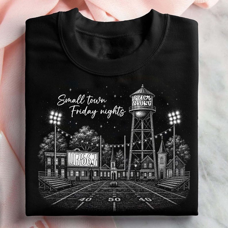 Op de afbeelding: Een zwart T-shirt met ronde hals en een witte grafische illustratie van een voetbalveld in de nacht. Het ontwerp bevat een watertoren met het opschrift "Petersburg", stadionverlichting en de tekst "Small town Friday nights" met "47567" op het scorebord.