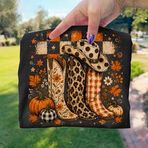 Puede incluir: Una camiseta negra con un estampado gráfico de tres botas vaqueras y un sombrero de vaquero. Las botas y el sombrero muestran patrones florales, de leopardo y de cuadros. El diseño incluye calabazas naranjas, hojas de otoño y estrellas sobre un fondo a cuadros.