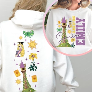 May include: A white hoodie and sweatshirt featuring a graphic print of a tower, a character with long blonde hair wearing a graduation cap, a green chameleon, yellow suns, lanterns, and a flower. The text reads Senior Class of 2027 Emily.