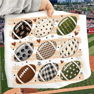 May include: A white t-shirt featuring a graphic print of nine American footballs in various patterns, including black, olive green, plaid, and star prints, arranged in rows against a background of horizontal tan brushstrokes and small hearts.