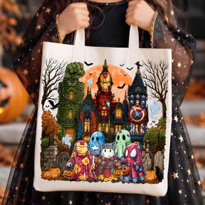 May include: A white canvas tote bag featuring a colorful Halloween-themed graphic of a haunted mansion with Iron Man, Captain America, Thor, and Spider-Man characters dressed in ghost costumes, surrounded by pumpkins, bats, and bare trees.
