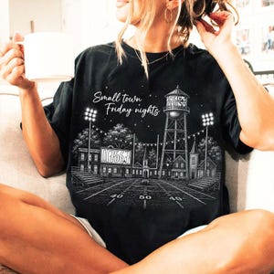 Op de afbeelding: Een zwart t-shirt met een witte grafische illustratie van een voetbalveld bij nacht, inclusief een watertoren, stadionverlichting en de tekst "Small town Friday nights" en "Petersburg" gedrukt boven het veld en op de watertoren.