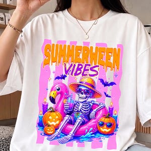 Könnte beinhalten: Weißes T-Shirt mit einem farbenfrohen Grafikdesign. Das Design zeigt ein Skelett mit Sonnenbrille und Hut, das auf einem rosa Flamingo-Schwimmer entspannt. Der Text "SUMMERWEEN VIBES" befindet sich über dem Skelett. Kürbisse, Kerzen und Fledermäuse ergänzen das Design.