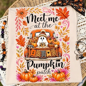 May include: A cream-colored t-shirt featuring a graphic of an orange pickup truck filled with pumpkins and a ghost wearing a beanie. The text reads Meet me at the Pumpkin patch. The design is surrounded by autumn leaves, flowers, and candy corn.