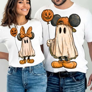 May include: Two white t-shirts featuring graphic prints of ghosts dressed as cartoon mice. The left shirt shows a ghost with a polka-dot bow and orange shoes holding a pumpkin balloon. The right shirt shows a ghost in a baseball cap holding a pumpkin balloon.