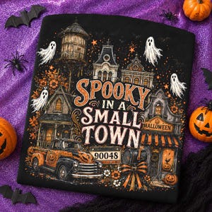 May include: A black t-shirt featuring a graphic print of a small town scene with a water tower, houses, a vintage truck, and ghosts. The text reads "SPOOKY IN A SMALL TOWN" and "90048" with orange and black autumn leaves and pumpkins throughout.