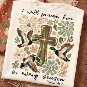 Peut inclure: Un t-shirt couleur crème orné d'une croix brune centrale entourée d'illustrations de trois canards colverts en vol et de fleurs aux tons sourds de vert, brun et beige. Le texte indique, I will praise him in every season PSALM 146:2.